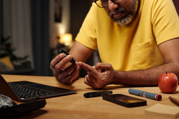 Man testing glucose level at home with medical device and laptop