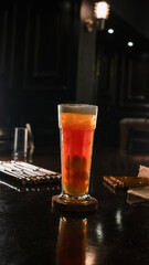 A tall glass of iced lychee tea with a vibrant orange gradient, containing whole lychee fruits at the base, served on a wooden coaster over a dark reflective surface, creating a refreshing and tropica