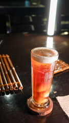 A tall glass of iced lychee tea with a vibrant orange gradient, containing whole lychee fruits at the base, served on a wooden coaster over a dark reflective surface, creating a refreshing and tropica