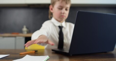 Boy in white shirt and tie focused on typing on laptop keyboard and writing on papers with pen. Shallow depth of field keeps actions clear indicating diligent work
