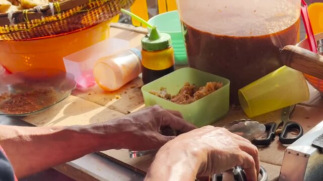 Street food seller preparing Batagor, a popular Indonesian fried dumpling with peanut sauce, using scissors and tongs at a traditional food stall in daylight