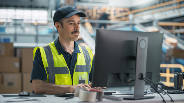 Male Senior Manager Checking Inventory On Desktop Computer In Warehouse Facility With Automated Conveyor Belt. Sorting Center Employees Carrying Boxes to Package Products And Shipping to Clients.