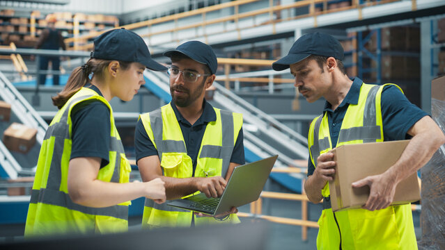 Diverse Team Of Warehouse Workers Having A Meeting In Logistics Facility With Conveyor Belt. Manager Using Laptop and Talking To Supervisor And Stocking Associate, Discussing Delivery Of Orders.