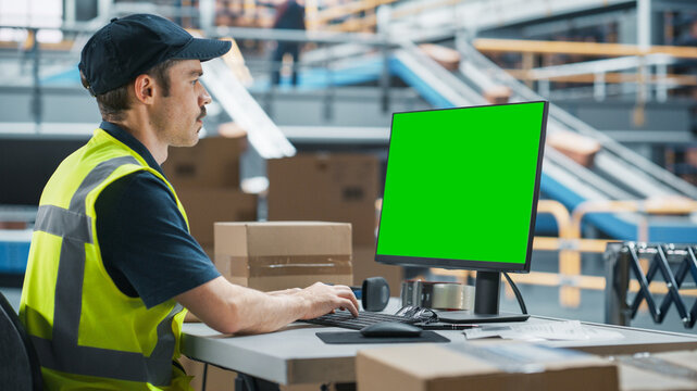 Caucasian Male Stocking Associate Using Green Screen Chromakey Desktop Computer In Distribution Facility. Multiethnic Male Warehouse Worker Loading Cardboard Boxes On Automated Conveyor Belt.