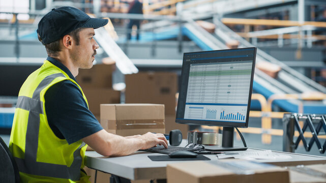Male Stocking Associate Checking Inventory On Desktop Computer In Warehouse Facility With Automated Conveyor Belt. Sorting Center Employees Carrying Boxes to Package Products, Deliver to Clients. - Powered by Adobe
