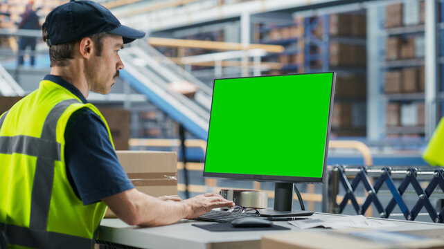 Caucasian Male Stocking Associate Using Green Screen Chromakey Desktop Computer In Modern Distribution Facility. Diverse Employees Working With Online Orders, Loading Cardboard Boxes On Conveyor Belt.