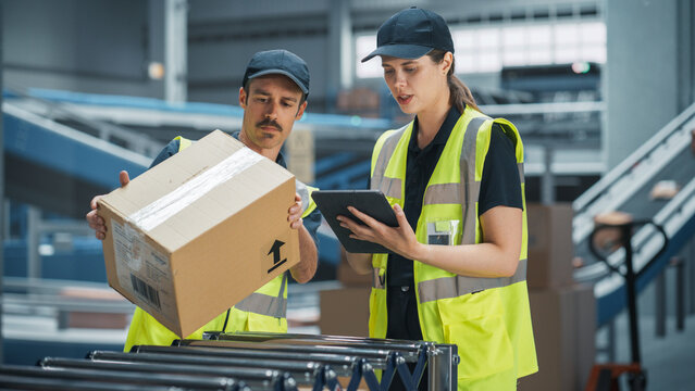 Caucasian Female Manager And Male Load Worker Talking In Warehouse Facility Of Logistics Company. Man Unloading Boxes From Automated Conveyor Belt, Woman Using Tablet Computer To Check Inventory. - Powered by Adobe