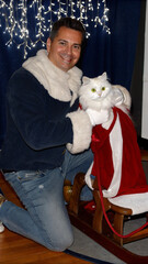 Obraz premium A close-up shot of Ded Moroz kneeling beside a fluffy white cat wrapped in a cozy red scarf. The cat sits on a wooden sleigh filled with wrapped gifts, its green eyes gleaming with curiosity. Ded Moro