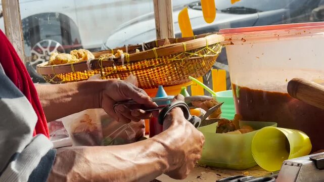 Street food seller preparing Batagor, a popular Indonesian fried dumpling with peanut sauce, using scissors and tongs at a traditional food stall in daylight