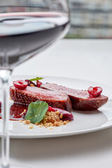 A gourmet dish of perfectly cooked duck breast with cherries, a rich sauce, and crumble, paired with a glass of red wine in the foreground.