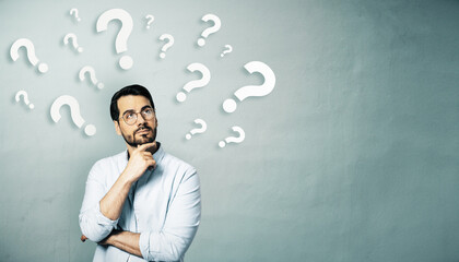 Thoughtful businessman standing in front of a wall with floating question marks, symbolizing...