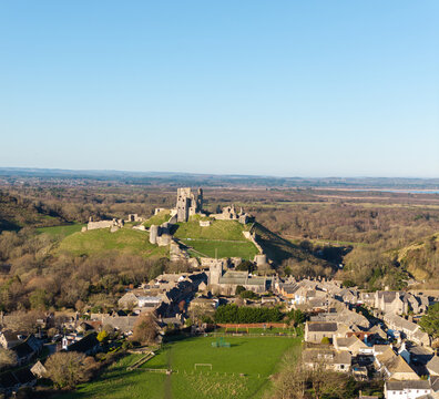 Aerial view of the medieval Corfe Castle ruins atop a grassy hill, overlooking the quaint village below, under a clear blue sky, Corfe Castle, England.