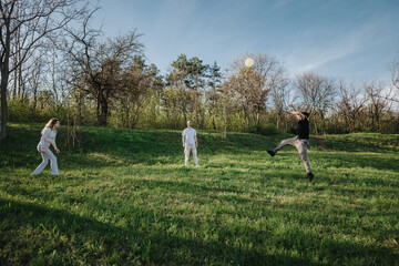 A group of three people engaging in playful activities outdoors on a sunny day, surrounded by nature and trees in a park-like setting, promoting bonding, fun, relaxation, and physical activity.