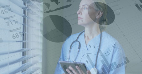 Clinician wearing scrubs with stethoscope, studying data charts on tablet by blinds in clinic