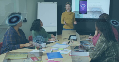 Presenting mid adult woman gesturing at conference table in office, with laptops, projection screen