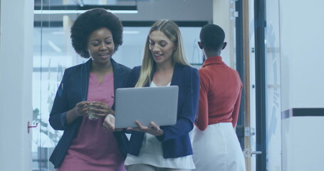 Two women walking and examining laptop in corridor with frosted partitions, holding glass of water