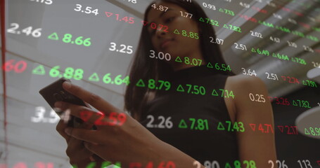 Businesswoman in sleeveless black top holding smartphone, reading stock figures on glass in office