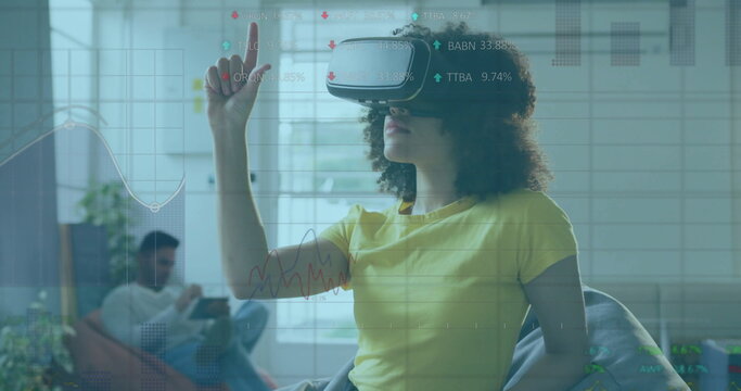 Pointing African American woman wearing VR headset interacting with virtual charts in living room