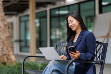 Asian businesswoman using laptop and smartphone sitting on bench outside office building