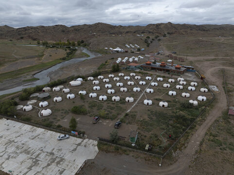 Aerial view of a yurt camp nestled in a rugged landscape where the arid earth meets the winding river under a vast, clouded sky, Turgen, Uvs, Mongolia.