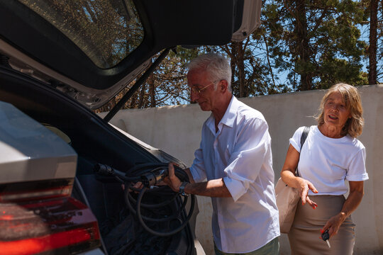 Senior man taking charging cable from car trunk, woman holding car key
