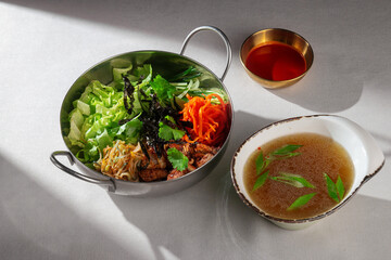Colorful Korean bibimbap without egg in a metal bowl, featuring fresh vegetables, grilled beef, cilantro, shredded seaweed and bean sprouts, served with a clear broth and gochujang sauce