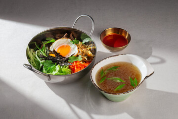 Authentic Korean bibimbap in a metal bowl with crispy breaded chicken, sunny side up egg, fresh vegetables, sprouts and seaweed, served with clear broth and spicy gochujang sauce