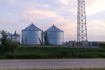 An industrial or agricultural setting, featuring several large grain silos under a vast sky at what appears to be either dawn or