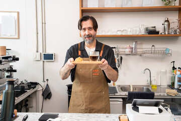 Smiling senior male café owner wearing a brown apron presents a tray with a cup of coffee and a pastry, standing confidently behind the counter in his small business, ready to serve customers.