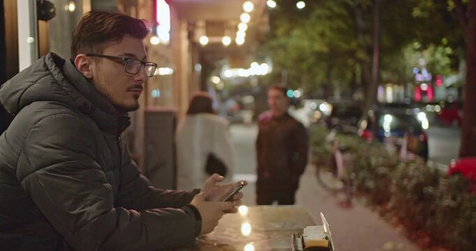 Young man sitting alone at table in a street coffee bar at night, copyspace.