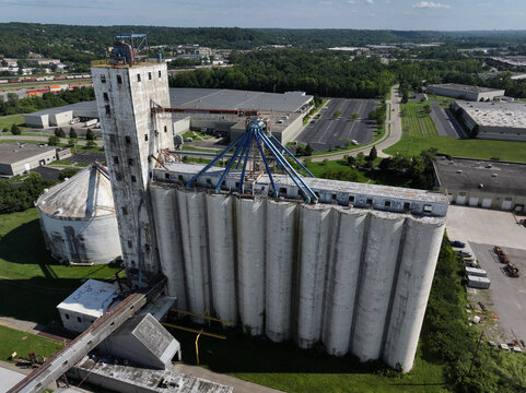 Aerial view of a towering grain elevator complex casting long shadows on the asphalt below, contrasting with the verdant trees in the distance, Cincinnati, Ohio, United States.