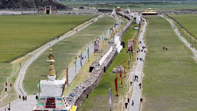 Aerial view of the Bage Mani Stone Wall, a long stone structure with stupas and flags, set against a backdrop of lush green fields, Bage, Sichuan, China.
