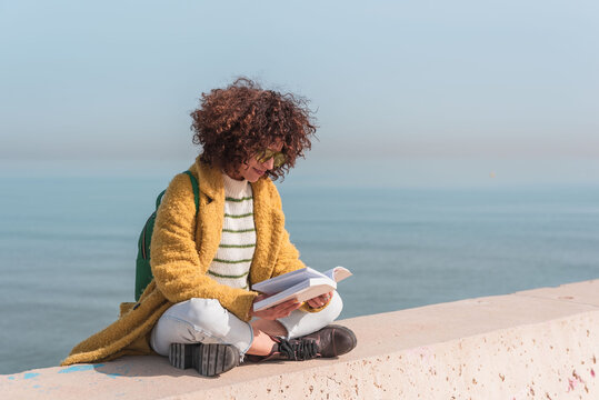 Young woman sitting with cross legs enjoying a relaxing day reading a book by the urban sea - Powered by Adobe