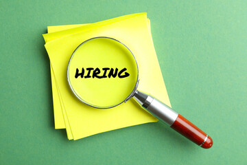 Magnifying glass on note paper with word Hiring and on green background, top view.