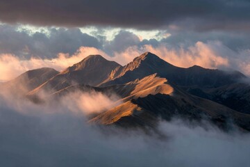 Fototapeta premium Dramatic mountain range at sunrise with low clouds, mist, and warm light across jagged peaks.