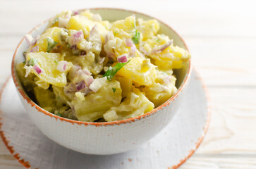 Cold potato salad with purple onion, gherkins and parsley in a white bowl on the table. Traditional German dish concept. Selective focus. Horizontal orientation. copy space.