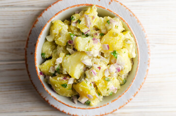 Cold potato salad with purple onion, gherkins and parsley in a white bowl on the table. Traditional German dish concept. Selective focus. Horizontal orientation. Top view