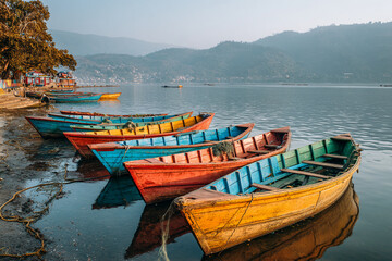 Obraz premium Row of colorful fishing boats moored along a quiet riverbank, reflection shimmering in the water, distant hills covered in morning mist