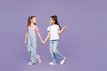 Full body smiling fun little two friends kids children girls 9-10 years old wear casual clothes together hold hands walk go point finger aside isolated on plain purple background. Lifestyle concept.