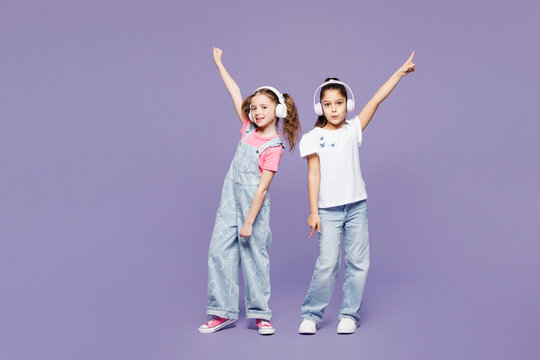 Full body fun little cheerful two friends kids children girls 9-10 year old wearing casual clothes together listen to music in headphones dance isolated on plain purple background. Lifestyle concept.