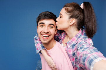 Close up young couple two friends family man woman wear pink casual clothes together doing selfie shot pov on mobile cell phone sit on back kiss isolated on plain blue background. Lifestyle concept.