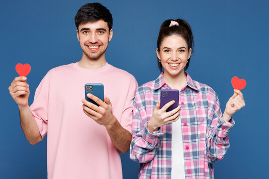 Young couple two friends family man woman they wearing pink casual clothes together use mobile cell phone using dating app hold paper heart isolated on plain blue background studio. Lifestyle concept