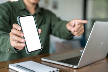 A man is holding a smartphone with a blank white screen and pointing at it while working on a laptop, showcasing a mobile app or website design