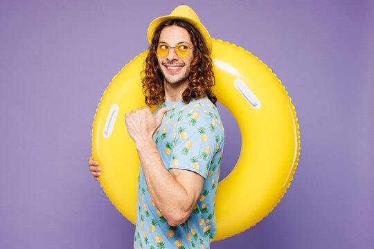 Young man wear blue beach t-shirt yellow hat sunglasses rest near hotel pool hold rubber ring point thumb finger aside on area isolated on plain purple background. Summer vacation sea sun tan concept. - Powered by Adobe