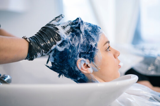 A young woman receives a hair wash from a gloved stylist at a salon. Her vibrant blue hair is covered in rich shampoo foam, indicating a professional hair treatment.