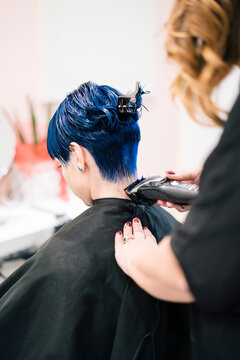 A professional hairdresser trims the vibrant blue hair of a client using an electric clipper. The stylist focuses on the back of the neck, providing a precise cut in a modern salon setting.
