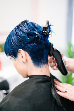 A person with striking blue, wet hair receives a professional haircut from a stylist using clippers. The hairdresser's hands are visible, adjusting the cape and trimming the nape.