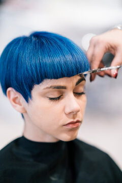 A person with vibrant blue hair receives a haircut at a salon. A stylist's hands carefully trim the bangs, focusing on precision. The client's eyes are closed.