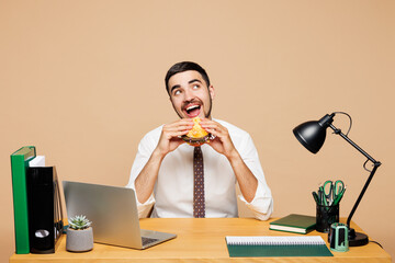 Young happy successful employee business man wear white shirt tie sit work at office desk with pc laptop eat fast food burger look aside isolated on plain beige background. Achievement career concept.