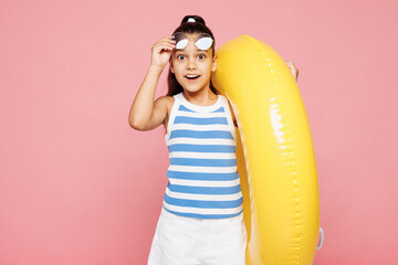 Traveler happy kid child wearing blue casual clothes rest near hotel pool hold inflatable rubber yellow ring take off sunglasses isolated on plain pink background. Summer vacation sea sun tan concept.